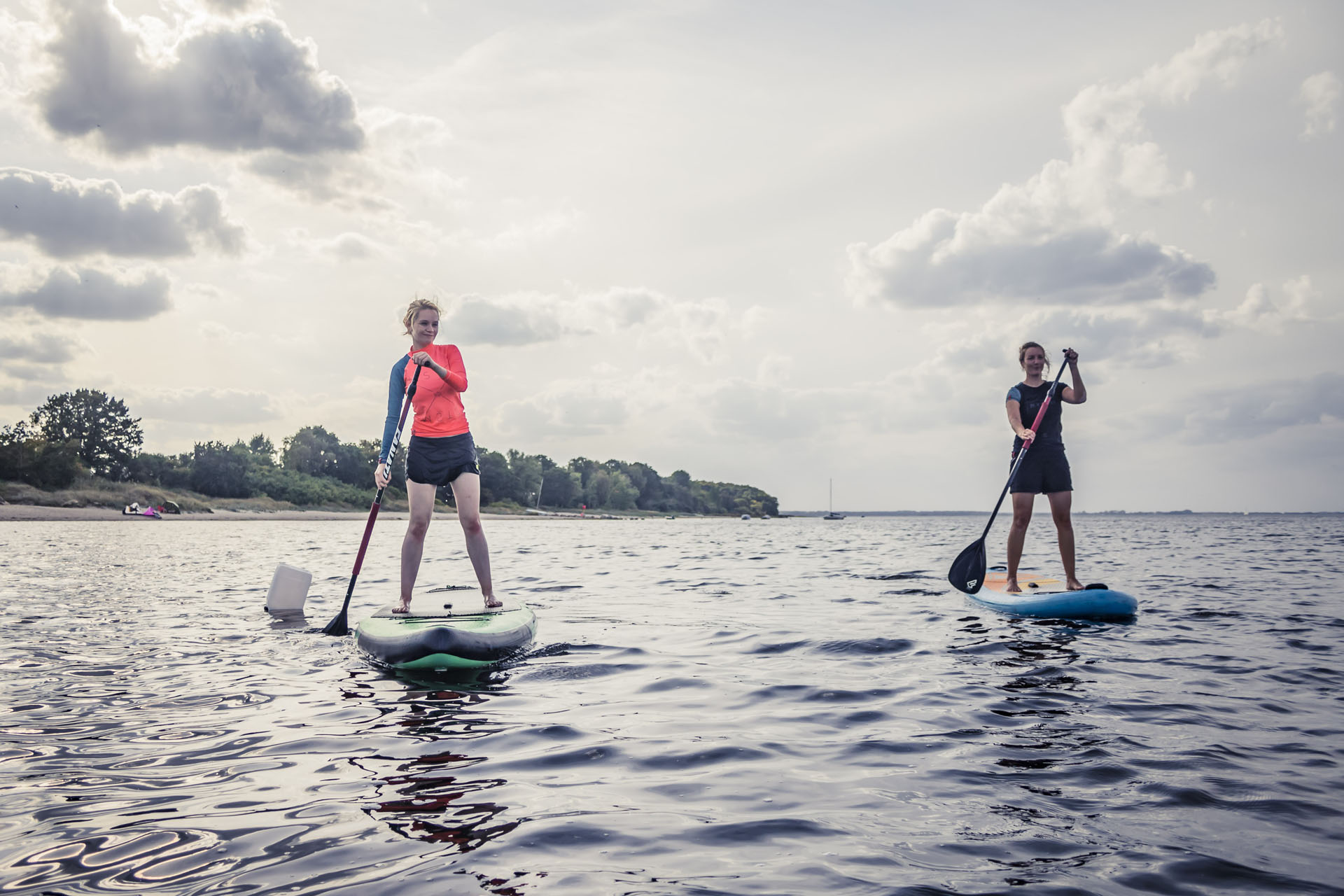 Zwei Frauen auf einem grünen und einem blauen SUP, eine links und eine rechts im Bild paddeln vom Naturstrand weg und um eine kleine Boje..